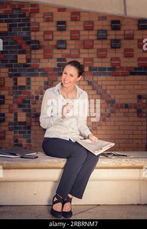 Attractive woman sitting on a wall bench laughing as she holds a notepad and paperwork in her hand that she was busy working on, decorative patterned Stock Photo