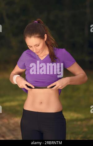 Woman athlete checking her pulse belt during a training run through woodland lifting her top to expose her toned slender midriff Stock Photo