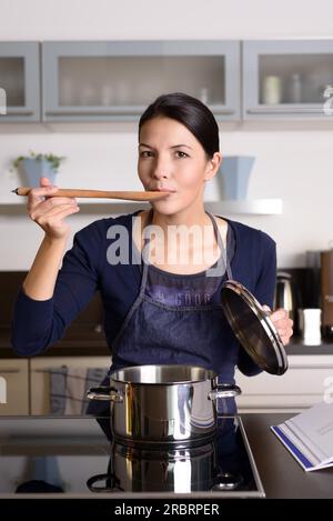 Young housewife tasting her cooking as she prepares the dinner in her kitchen sampling directly from the pot on the hob with a wooden spoon as she Stock Photo