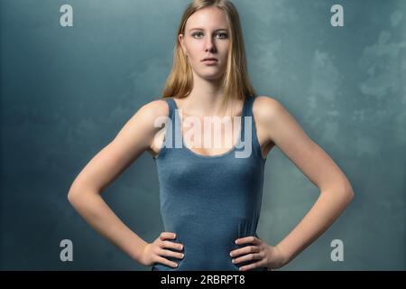 Serious attractive young woman with an attitude standing akimbo with her hands on her hips staring intently at the camera against a green chalkboard Stock Photo