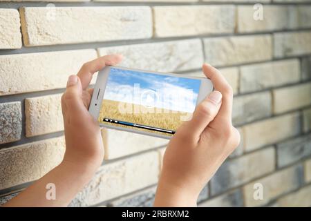 Woman watching video on mobile phone near brick wall indoors, closeup Stock Photo