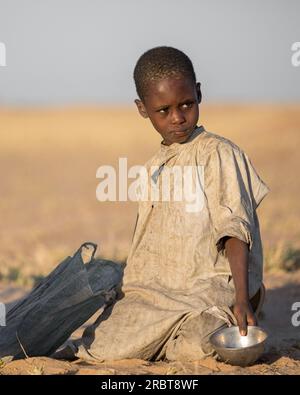 Young boy, clad in a weathered robe, humbly kneels on the steppe ground, holding a small metal bowl in his left hand, echoing the resilience of life Stock Photo