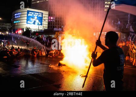 July 6, 2023, Tel Aviv, Israel: Israeli anti-reform protesters wave the Israeli flag next to a bonfire on the blocked Ayalon highway as they are sprayed with water from police water cannons during the demonstration. Thousands of Israelis took to the streets and blocked the Ayalon highway in Tel Aviv clashing with police who used water cannons and mounted police officers and arrests to disperse the protesters after Tel Aviv police chief Amichai Eshed announced his resignation from the force. (Credit Image: © Matan Golan/SOPA Images via ZUMA Press Wire) EDITORIAL USAGE ONLY! Not for Commercial U Stock Photo