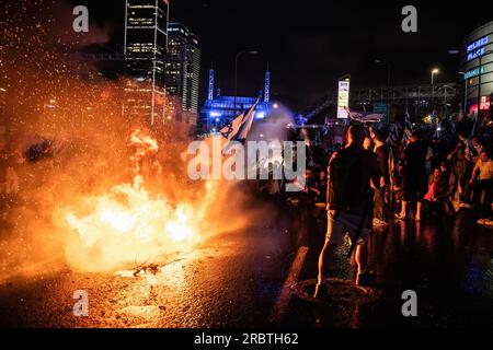 July 6, 2023, Tel Aviv, Israel: Israeli anti-reform protesters wave the Israeli flag next to a bonfire on the blocked Ayalon highway as they are sprayed with water from police water cannons during the demonstration. Thousands of Israelis took to the streets and blocked the Ayalon highway in Tel Aviv clashing with police who used water cannons and mounted police officers and arrests to disperse the protesters after Tel Aviv police chief Amichai Eshed announced his resignation from the force. (Credit Image: © Matan Golan/SOPA Images via ZUMA Press Wire) EDITORIAL USAGE ONLY! Not for Commercial U Stock Photo