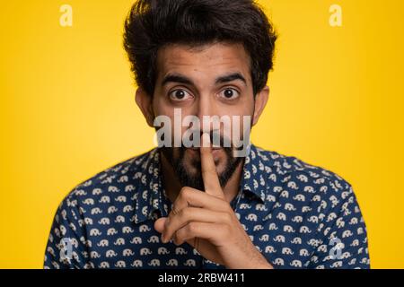 Shh be quiet please. Young indian man presses index finger to lips makes silence gesture sign do not tells secret, stop gossip. Handsome bearded hindu guy isolated alone on yellow studio background Stock Photo