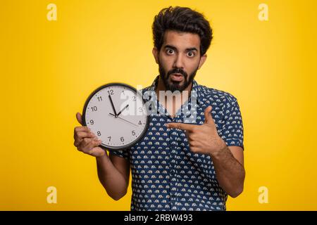 Confused indian man with anxiety checking time on clock, running late to work, being in delay deadline. Hindu guy looking at hour, minutes, worrying to be punctual isolated on studio yellow background Stock Photo
