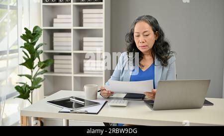 A serious and focused Asian senior businesswoman or female boss reading a business report at her desk in her office. Stock Photo