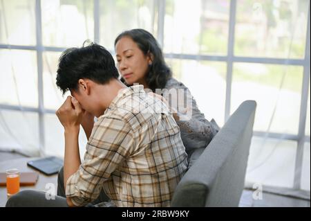 A caring Asian mom is trying to comfort and support her crying and sad son while sitting on a sofa together. care, family support, stress relief, bond Stock Photo