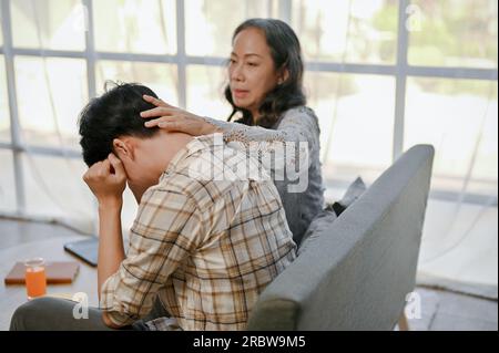 A caring Asian grandmother is sitting on a sofa with her sad grandson, trying to calm and comfort him from his stress and disappointment. Stock Photo