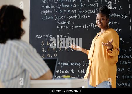 African American tutor standing near the blackboard and explaining IT material to student at lesson Stock Photo