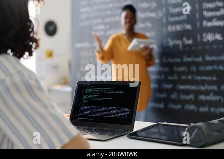 Rear view of student typing codes on computer while sitting at desk at lesson with teacher explaining material in background Stock Photo