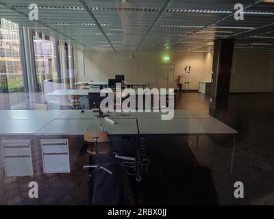 Empty office space with desks, chairs, computer monitors. Sunlight coming in from large window walls on the side, no people, seen from outside. Stock Photo