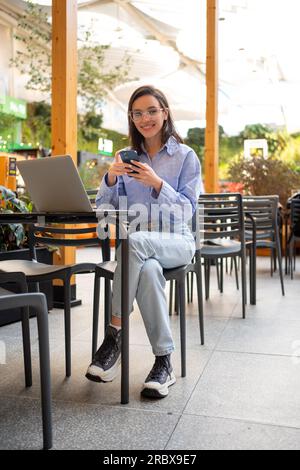 Freelance worker doing remote work using smartphone and laptop, looking at camera and smile. Female freelancer with mobile phone in outdoor cafe. Full length vertical shoot Stock Photo