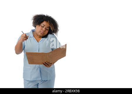 young afro female doctor wearing a blue uniform, speaking on a phone call taking notes on a brown folder with a pen, white background with copy space. Stock Photo
