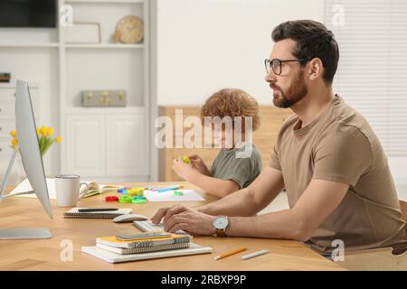 Man working remotely at home. Father using computer while his son playing at desk Stock Photo