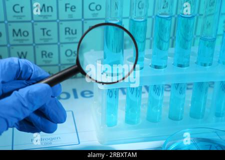 Scientist with magnifying glass examining test tube against periodic table of elements in laboratory, closeup. Color tone effect Stock Photo