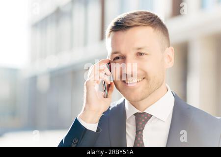 Confident happy middle aged businessman communicates via cell phone, discusses financial report with business partner, wears formal clothes, stands ou Stock Photo