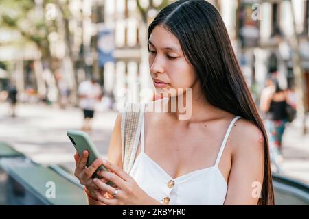 Latin American female reading news on cellphone on blurred background of sunlit street in Barcelona, Spain Stock Photo