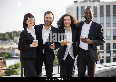 Portrait of friendly multiethic team of colleagues posing together and looking at camera. Four young cheerful people with coffee and gadgets smiling and having fun while meeting at splendid terrace. Stock Photo