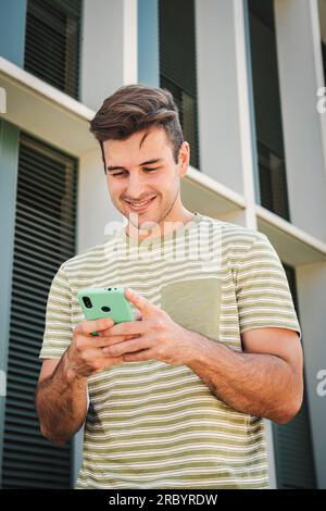 Vertical portrait of happy teenager male smiling and typing on a smart phone using a social media app outdoors. Young man having fun texting messages on a cellphone. Handsome guy using a mobile phone . High quality photo Stock Photo