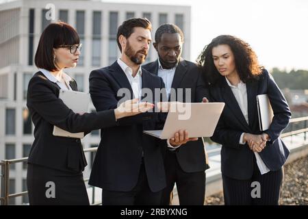 Close up of diverse successful employees from sales department watching online presentation on portable laptop outdoors. Caucasian colleagues pointing with hand at screen and analyzing numbers. Stock Photo