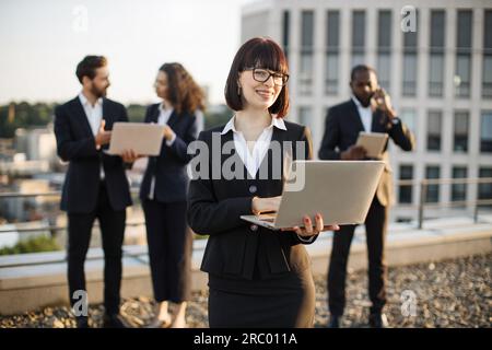 Portrait of charming business lady with short dark hair holding portable laptop and making frank smile on camera. Handsome african american man talking on cellphone while his partners disputing. Stock Photo