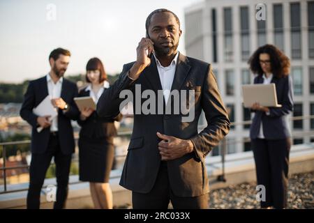 Beaming african american gentleman having conversation on smartphone during meeting with partners on terrace. Multiracial colleagues in suits using re Stock Photo
