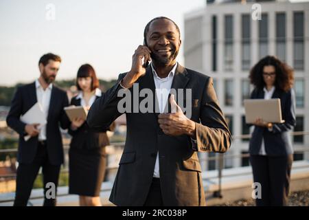 Beaming african american gentleman having conversation on smartphone during meeting with partners on terrace. Multiracial colleagues in suits using re Stock Photo