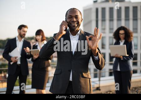 Beaming african american gentleman having conversation on smartphone during meeting with partners on terrace. Multiracial colleagues in suits using re Stock Photo