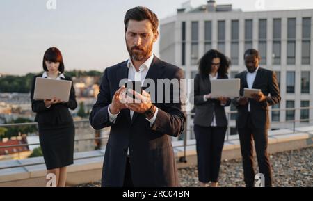 Caucasian bearded entrepreneur dialing number on smartphone while his business partners using portable devices on background. Multiracial investors working together on plan of creation new complex. Stock Photo