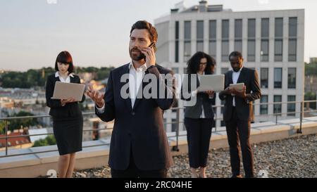 Business investor talking on smartphone while his colleagues working on personal gadgets. Multicultural partners in formal attire using modern technologies for planning new architectural project. Stock Photo