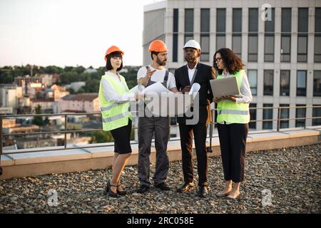 Team of multiracial architects and designers standing on panoramic terrace and looking together at project schematics. African american lady holding r Stock Photo