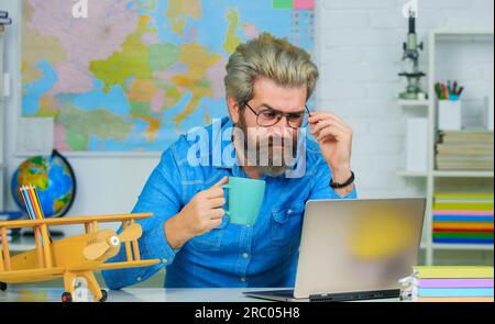 Serious male teacher drinking coffee or tea in classroom. Teacher or university professor at workplace. School teacher in glasses sitting at desk in Stock Photo