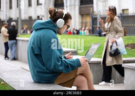 Guy in shorts and headphone sitting with a laptop on a bench on summer street Stock Photo