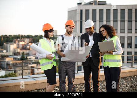 Group of four multiracial people in suits and helmets brainstorming for common project of new real estate building. Construction team working with blueprints and modern laptop in fresh air. Stock Photo