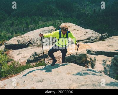 Ginger hair man tourist wear bright yellow jersey in mountain trail, walking on sandstone rocky hill, wearing backpack and sunglasses, using trekking Stock Photo