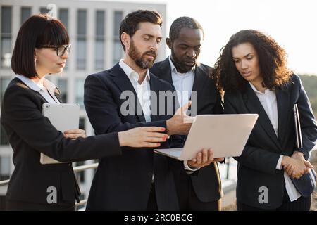 Close up of diverse successful employees from sales department watching online presentation on portable laptop outdoors. Caucasian colleagues pointing with hand at screen and analyzing numbers. Stock Photo