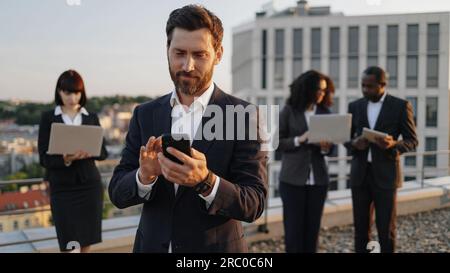 Caucasian bearded entrepreneur dialing number on smartphone while his business partners using portable devices on background. Multiracial investors working together on plan of creation new complex. Stock Photo