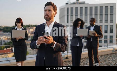 Caucasian bearded entrepreneur dialing number on smartphone while his business partners using portable devices on background. Multiracial investors working together on plan of creation new complex. Stock Photo