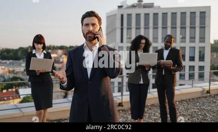 Business investor talking on smartphone while his colleagues working on personal gadgets. Multicultural partners in formal attire using modern technologies for planning new architectural project. Stock Photo