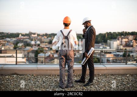 Back view of two male developers wearing hardhats standing on rooftop and talking about apartment facades. Workman in building uniform holding blueprints while business man touching chin and thinking. Stock Photo