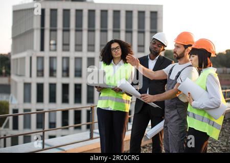 Four multiracial engineers with technical drawings and laptop discussing place for construction of new multi-apartment complex. Workman talking and pointing with hand while others listening carefully. Stock Photo