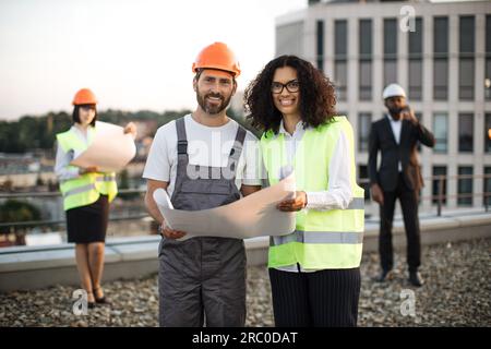 Portrait of caucasian foreman in hardhat and african american inspector in vest looking at camera and smiling on roof. Businessman in suit talking on smartphone while his colleague looking at plan. Stock Photo