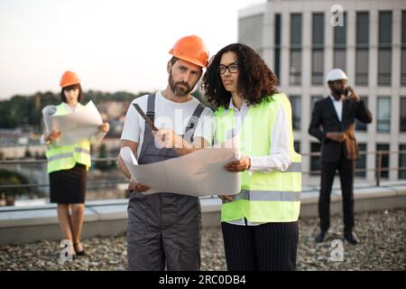 Foreman with ruler in hands talking to inspector and explaining designing presented on paper architectural plan. Male investor in suit talking on smartphone while his colleague watching at blueprint. Stock Photo