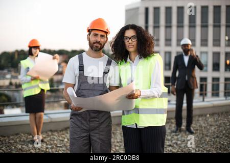 Portrait of caucasian foreman in hardhat and african american inspector in vest looking at camera and smiling on roof. Businessman in suit talking on smartphone while his colleague looking at plan. Stock Photo