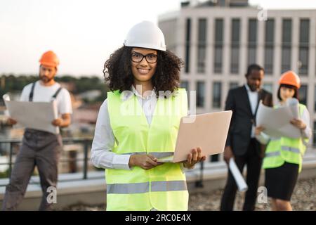 Portrait of african american female urban planner in protective wear and glasses holding remote computer and looking at camera. Diverse architects reviewing building blueprints on roof terrace. Stock Photo