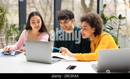 Happy international students friends sitting at desk in coworking space and using laptop, making project together Stock Photo