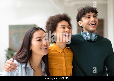 Happy diverse friends embracing looking aside and smiling, enjoying break during classes, standing in university hallway Stock Photo