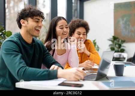 Happy three diverse friends studying together in coworking space, learning with laptop preparing for university exam Stock Photo