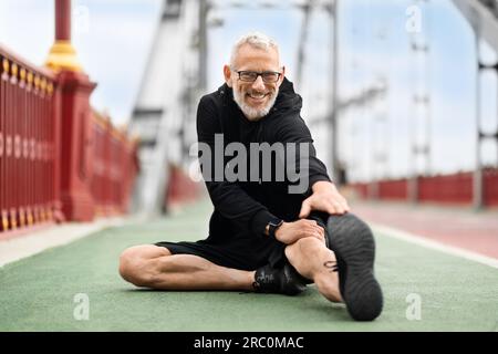 Healthy sporty senior man sitting on ground, stretching body Stock Photo